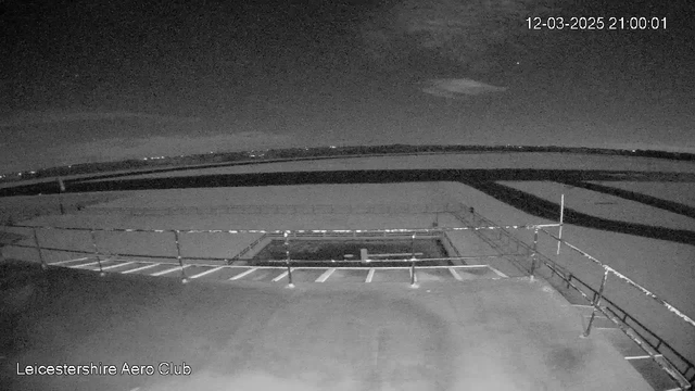 A black-and-white image showing a wide view of a dark landscape at night. In the foreground is a rooftop with a railing, and a rectangular area that appears to be a helipad marked with a white "H." The background features a flat surface, possibly water or land, with a dark horizon and some faint lights visible in the distance. The date and time are displayed in the upper right corner.