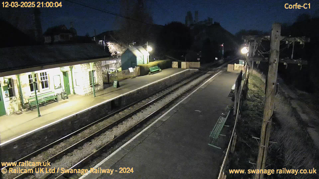 A railway station at night, featuring low lighting. The platform is empty with a few wooden green benches visible. A stone building with large windows is on the left side, indicating a waiting area. There is a green sign that reads "WAY OUT." Two parallel tracks run through the center, bordered by gravel. A pole with wires stands to the right, and a wooden fence encloses a grassy area at the far end of the platform. The atmosphere is quiet and dimly lit.