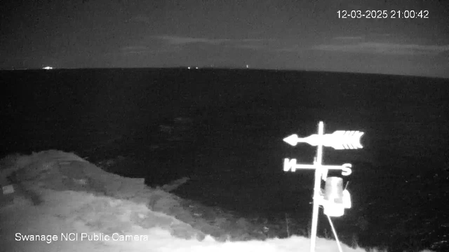 A dark scene showing the ocean at night, with faint lights visible on the horizon. In the foreground, a weather vane stands on a rocky shoreline, indicating directions labeled North, South, East, and West. The timestamp in the corner shows the date and time as December 3, 2025, at 21:00:42.