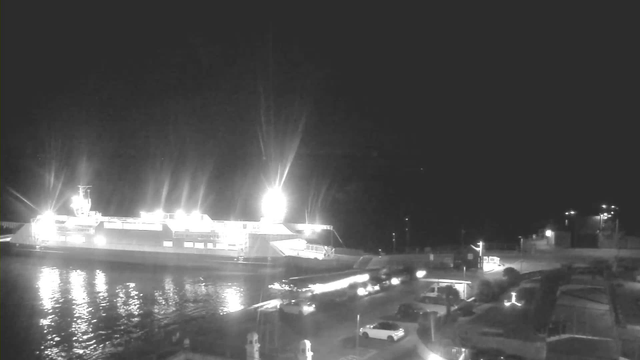 A night-time view of a harbor scene with a large ferry docked. The ferry is brightly illuminated with multiple lights, reflecting off the water. In the foreground, there is a parking area with several cars visible. The environment is mostly dark, but some streetlights can be seen in the background, along with a few structures. The overall mood is quiet and calm, typical of a late evening by the water.