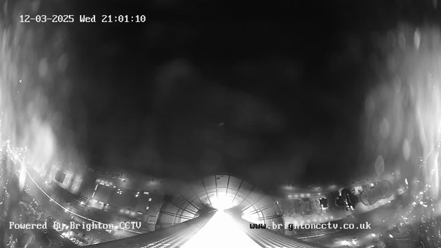 A night-time webcam view showing a brightly lit area at the center fading into darkness around the edges. The perspective suggests a view looking down a street or corridor, with blurred lights and outlines of structures visible. The image includes time and date at the top, indicating it was captured on December 3, 2025, at 9:01 PM. The logo of Brighton CCTV appears in the corner.