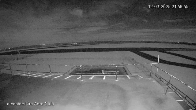 A nighttime view from a webcam at the Leicestershire Aero Club. The foreground features a flat, dark surface with a railing along the edge. In the background, there is a vast open area, possibly an airstrip, with faint lines indicating runway markings. The sky is mostly cloudy with dim lighting, contributing to a monochromatic color scheme. The date and time are displayed in the top right corner, indicating it is 21:59:55 on March 12, 2025.