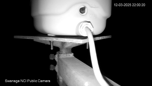 The image shows a dark scene featuring a white container resting on a metal plate. A power cord is connected to the container, and there are several metallic parts visible beneath it, including screws and a support arm. The background is mostly black, indicating low light conditions. In the corner, a timestamp displays "12-03-2025 22:00:20," with a label indicating it is from the "Swanage NCI Public Camera."