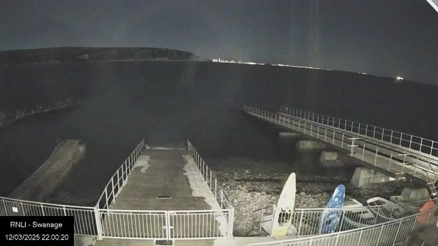 A nighttime scene showing a calm body of water with a rocky shoreline. In the foreground, there is a concrete pier leading to the water, with a walkway enclosed by a white railing. On the right side, there are two kayaks, one blue and one white, resting on the ground. The distant shoreline is faintly visible against the dark sky, with a faint line of lights indicating a built-up area. The image has a dark overall tone, suggesting a peaceful but quiet nighttime ambiance.