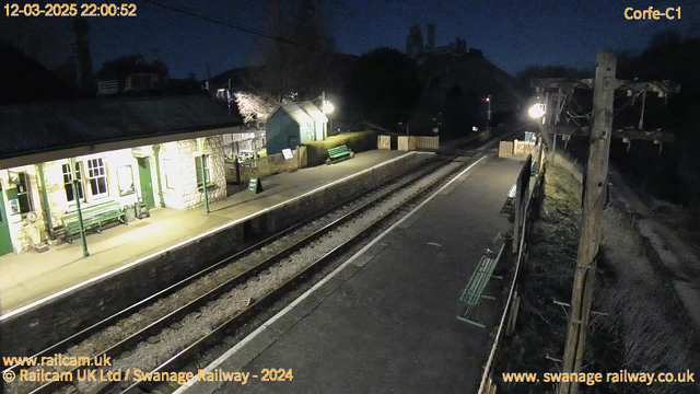 A dimly lit railway station is depicted at night. The foreground shows a platform with two sets of railway tracks. On the left, there is a stone building with large windows and green accents, along with a green bench underneath an awning. A sign reading "WAY OUT" is positioned near the building. The platform extends to the right where additional benches are visible. In the background, there are distant structures obscured by darkness, and a tall utility pole with a single light illuminates part of the scene. The overall atmosphere is quiet and serene.