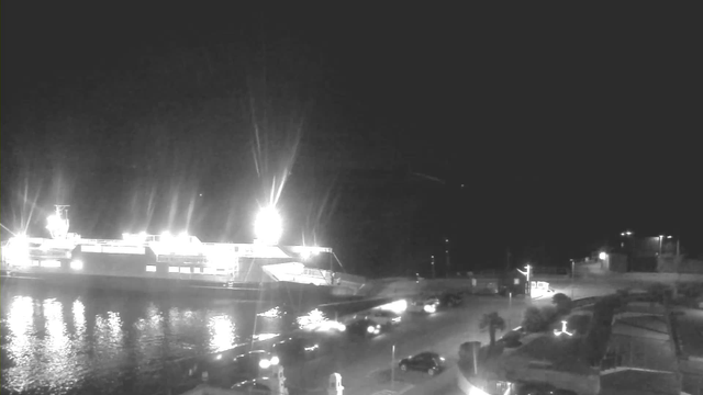 A brightly lit ferry is docked at night along a dark waterfront. The reflection of lights glimmers on the water surface. There are a few parked cars on the road beside the ferry, and some streetlights illuminate the area. The background is mostly dark, with hints of buildings and palm trees.