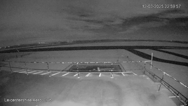 A dark, nighttime webcam image showing an elevated view of an airport area. In the foreground, a wooden railing surrounds a flat surface, partially obscuring white lines that likely mark parking spots. Beyond the railing, a wide, empty runway stretches out with a dark surface and a faint outline of another area. The sky is filled with clouds, and the overall scene is subdued with very little visible lighting, suggesting a quiet, nighttime atmosphere. The timestamp in the corner indicates it is 22:59:57 on March 12, 2025.