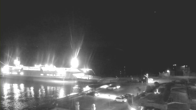 A ferry is docked at a harbor at night, illuminated by bright lights. The water reflects the lights, creating a shimmering effect. In the foreground, there are several parked cars and a paved area next to the water. The background features a dark sky with a few faint lights, possibly from buildings or other structures nearby.