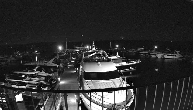 A nighttime scene at a marina, with several boats moored in a harbor. The image is in black and white, showing a row of yachts and smaller boats illuminated by dock lights. The wooden walkway between the boats is visible, leading to the larger vessel at the forefront. The background is dark, suggesting a calm evening with minimal activity.