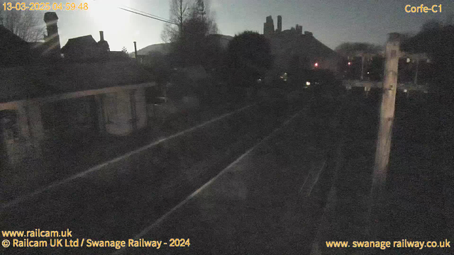 A dark webcam image showing a railway track at early dawn. On the left, there is a building with a chimney, partially illuminated by the low light. The background features a silhouette of hills and trees, with faint outlines of structures. There are overhead power lines, and a distant signal light is visible to the right. The timestamp at the top indicates the date and time of the image capture.