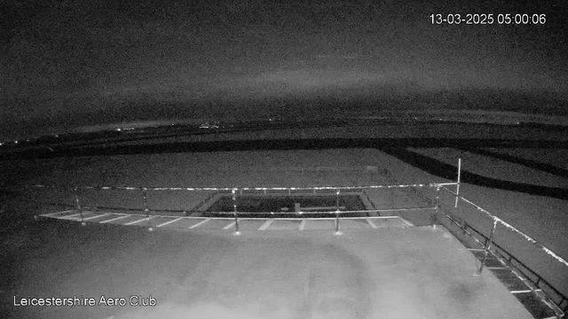 A dark, night-time scene viewed from a webcam at the Leicestershire Aero Club. The image shows a flat, empty rooftop area with a railing at the front. In the background, blurred lights can be seen on the horizon, indicating distant structures or vehicles. The surface of the rooftop appears mostly empty and smooth, and there is a rectangular dark area, possibly an opening or feature on the roof. The date and time in the corner are set to 13th March 2025 at 5:00 AM.