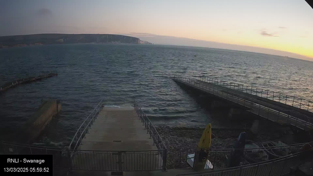 A view of the sea during dawn. The water is calm, with gentle waves lapping at the shore. In the foreground, there is a concrete ramp leading down to the water, surrounded by metal railings. To the right, a small jetty extends into the water. On the bank, there are various boats and equipment, including yellow and blue items. In the background, cliffs and a shoreline are visible against the morning sky, which shows subtle colors of orange and pink as the sun rises.