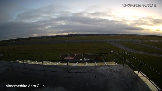 A view from a high vantage point at Leicestershire Aero Club, depicting a grassy airfield with runway markings. The sky is predominantly cloudy, with a soft light suggesting early morning. In the foreground, there's a railing with a flat rooftop surface visible, while the background showcases the airfield stretching into the distance, bordered by distant hills.