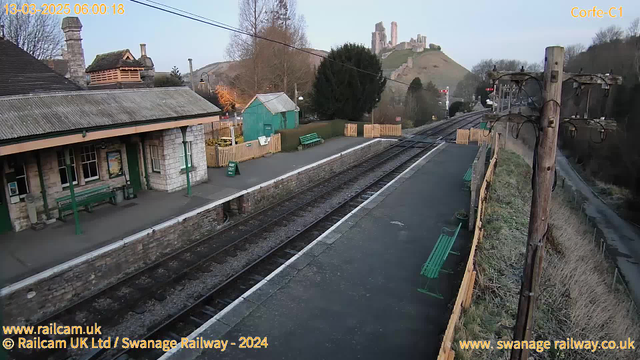 A railway station scene at dawn, featuring a stone building with a sloping roof and several windows. Green benches are positioned on the platform, and there's a wooden fence separating the station from a grassy area beyond. In the background, a hill rises with remnants of a castle at the top. Electrical poles are visible on the right side of the image, with overhead wires connected. The atmosphere is quiet and calm, indicative of early morning.