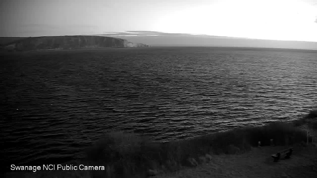 A black and white image showing a vast body of water with gentle waves. In the background, there are cliffs that rise above the shoreline, and the sky above is mostly overcast with soft clouds. On the lower part of the image, there is a grassy area with a couple of benches facing the water. The scene conveys a tranquil coastal landscape.