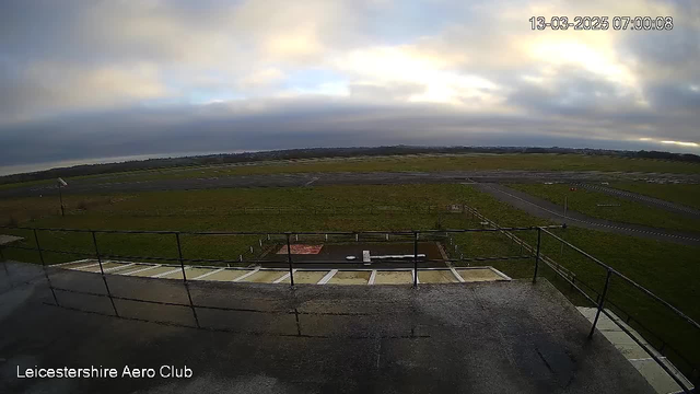 A webcam view overlooking an airfield. In the foreground, there is a railing and a cement platform. Beyond the railing, the airfield stretches out with a grassy area, some markings on the ground, and a light path in the distance. The sky is cloudy with varying shades of gray and blue. The date and time are displayed in the top right corner.