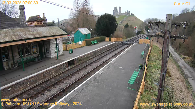 A railway station scene with stone and wooden structures. The station building has a sloped roof and a sign indicating the way out. There are benches along the platform, painted green, and a wooden fence surrounding the station. In the background, a hill rises featuring the ruins of a castle. The scene is set in a rural area with sparse trees and a cloudy sky. Tracks are visible, leading away from the platform. A utility pole stands on the side, equipped with wires.