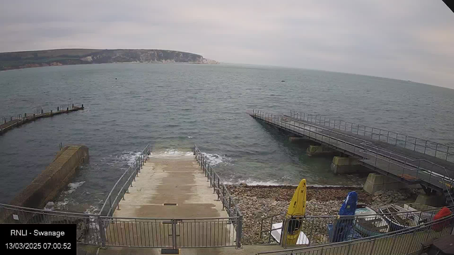 A view of a coastal area featuring a grey sky and calm sea. The shoreline has a series of steps leading down to the water, flanked by metal railings. On the right side, there is a small dock extending into the sea, with a yellow kayak and a blue kayak stored nearby. In the background, a cliff rises along the coastline. The image is timestamped at 07:00:52 on March 13, 2025.