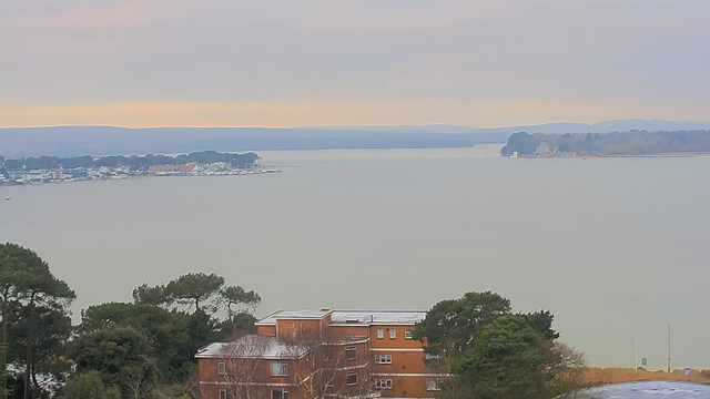 A landscape view showing a calm body of water with a slight fog. In the foreground, a brown brick building with a flat roof is visible, surrounded by green trees. The middle ground features small boats and the edge of a marina with various structures along the shore. In the background, rolling hills can be seen under a gray sky with soft, diffused light, indicating either early morning or late afternoon.