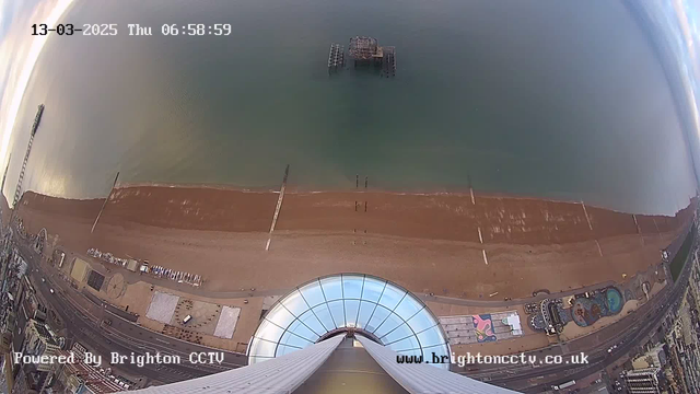 A view from a high vantage point showing a beach with golden sand and a calm sea. The shoreline is lined with wooden posts extending into the water. In the distance, there is a partially submerged pier structure. Below, there are colorful buildings and attractions, including a circular entrance with glass. The sky is cloudy with a soft light, indicating early morning. The date and time display is in the upper left corner: "13-03-2025 Thu 06:58:59."