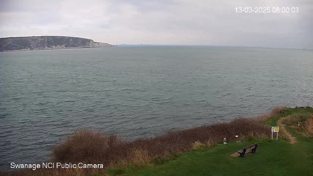 A view of a calm sea under a cloudy sky, with gentle waves rippling on the water's surface. In the foreground, there is a grassy area with some low bushes. A wooden bench is positioned to the right, and a sign is nearby. In the background, a landmass rises from the water, characterized by rugged cliffs, while the horizon stretches across the distance where the sea meets the sky. The overall atmosphere is tranquil and natural.