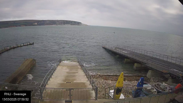 A view of the sea with gentle waves, under a cloudy sky. In the foreground, a set of stone steps leads down to the water's edge, flanked by railings. To the right, there are two piers extending into the water, one of which is longer and has a flat surface. Nearby, several brightly colored kayaks, including yellow and blue, are secured against a railing on a pebbly shore. The background features cliffs that rise above the coastline.