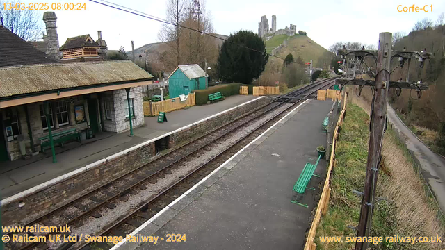 A railway station platform is shown, featuring a stone building with a sloped roof and several benches. There are tracks running along the platform, with a grassy hillside and a castle visible in the background. Green benches are located on the platform, alongside a sign that says "WAY OUT." The scene is set in a rural area, with trees and a road on the right side. The sky appears cloudy.