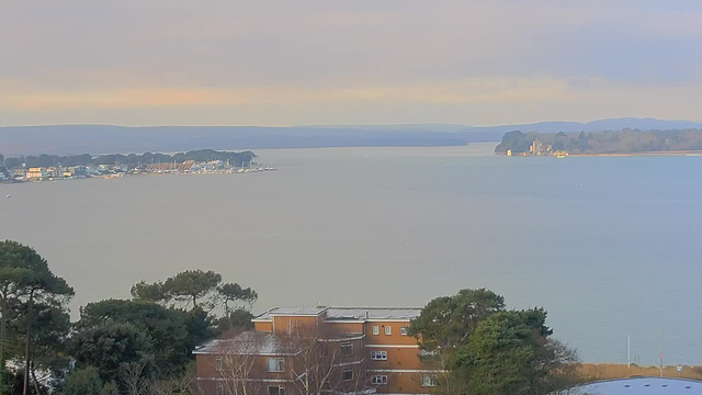 A serene view of a calm body of water stretching towards the horizon, with distant land and trees visible on both sides. In the foreground, there are some buildings surrounded by greenery. Small boats are scattered on the water, and soft, muted colors create a tranquil atmosphere in the sky and reflections on the water.