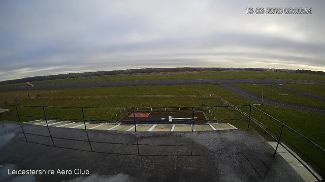 A view from a high vantage point over an airfield. The foreground features a railing of a balcony, and below is a grassy field with a few scattered fences. In the distance, the runway stretches across the image, which is mostly clear. The sky is overcast with subdued light, suggesting morning or early afternoon. The bottom left corner displays the text "Leicestershire Aero Club," with a timestamp in the top right corner indicating the date and time.