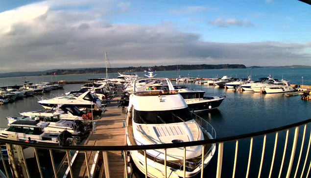 A dock filled with various boats and yachts, arranged closely together on calm waters. The scene is set under a partially cloudy sky, with some sunlight breaking through. In the background, a hilly shoreline is visible, partially covered by trees. The foreground features a wooden walkway leading to the boats, with a railing visible in the lower part of the image.