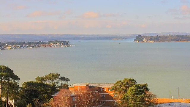 A view of a calm body of water with distant hills under a light blue sky. In the foreground, there are trees and a brick building with several windows. The shoreline is lined with houses and small boats, while in the background, green hills rise up. The overall scene conveys a peaceful, natural landscape.