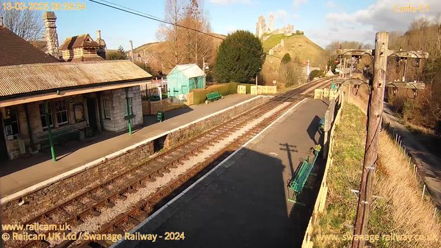 A railway station platform is depicted, showing multiple sets of railway tracks running parallel. There is a stone platform with a brick edge, benches painted green, and signs indicating paths. In the background, a hill rises with ruins of a castle or fortification on top, surrounded by trees. The sky is clear with some clouds, indicating a bright day. On the left, there is an older building with a sloped roof and a chimney, while a small blue building is visible in the middle ground. A power pole with wires is present on the right side of the image.