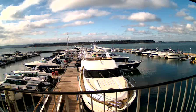 A marina scene featuring numerous boats docked in a calm body of water. The foreground shows a wooden walkway lined with boats of various sizes, including a large, white motor yacht. The background displays a tranquil landscape with distant hills under a partly cloudy sky, and a few scattered clouds. Sunlight reflects off the water's surface, contributing to the serene atmosphere.