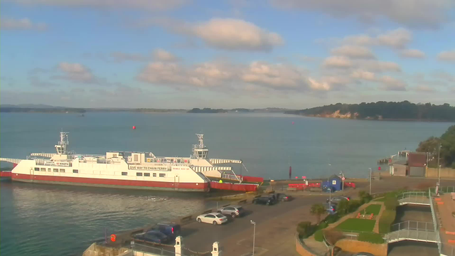 A white ferry with red trim is docked near a pier at a calm waterway, surrounded by rolling hills in the background. The sky is partly cloudy with blue patches visible. There are several parked cars in a lot beside the ferry, and some structures including a blue building and a dock can be seen along the shore.