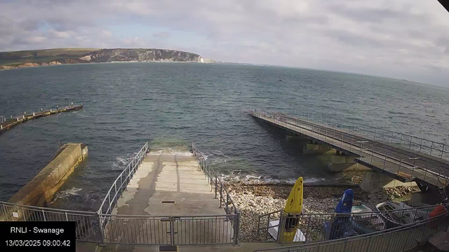 A view of a coastline with a rocky shore and a calm sea. In the foreground, there is a concrete ramp leading down to the water, flanked by a metal railing. Two piers extend into the water, with one curving to the left and another straight ahead. On the right side, a few boats are visible, including a yellow kayak and a blue vessel. The background features green hills and cliffs under a partly cloudy sky.