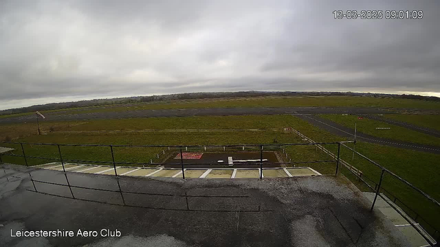 A cloudy sky is visible above an open field and runway at the Leicestershire Aero Club. The area is flat and green, with a paved runway and some white markings. In the foreground, a fenced area is present, and the scene appears to be early morning, with no visible aircraft or people.