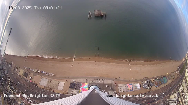 An aerial view of a sandy beach along the coastline. The beach is bordered by the sea, with gentle waves lapping at the shore. A large, abandoned pier structure is visible in the water. Various people can be seen walking on the beach, with paths leading towards the water. To the right of the image, there are buildings along the promenade, including colorful shapes and designs on the ground. The sky is partially cloudy with patches of blue. The bottom of the image features part of a building or structure, reaching upwards into the frame.