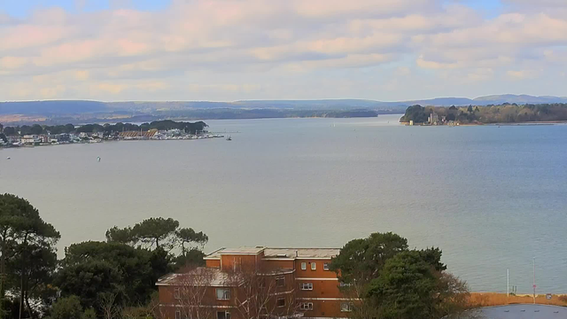 A panoramic view of a calm bay featuring varying shades of blue water under a cloudy sky. In the foreground, there are scattered green trees and a reddish-brown building. The shoreline opposite includes a marina with boats, while the distant background shows rolling hills with patches of green vegetation. The overall atmosphere is serene and natural.