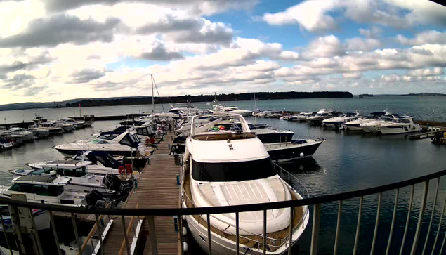 A view of a marina filled with various boats and yachts moored along a wooden dock. The water is calm and reflects the clouds in the sky, which are fluffy and white, scattered above a distant green shoreline. The scene is framed by a railing in the foreground, suggesting a viewpoint from an elevated position.