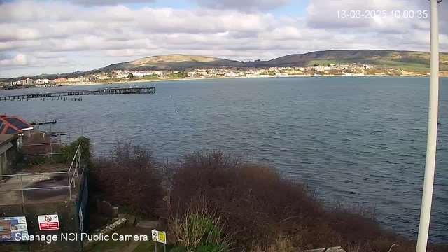 A coastal scene showing calm waters with a gentle shoreline. In the foreground, there are some bushes and a partially visible structure with a red roof. Further out, a wooden pier extends into the water. The background features a town with buildings along the coast, and hills rise up behind them. The sky is partly cloudy.