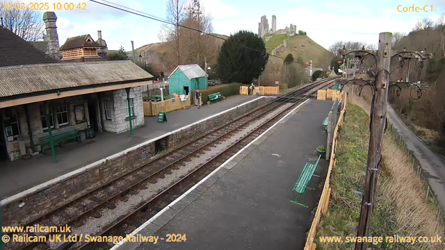 A railway station scene with two sets of tracks running horizontally across the image. On the left side, there is a stone building with a sloped roof, featuring a green bench in front and a small blue shed nearby. A few people are standing on the platform, which is bordered by a wooden fence. In the background, a hill rises to the right with ruins of a castle visible at the top, and trees are scattered throughout the area. The sky is partly cloudy, and a power pole with wires stands on the right side of the image.