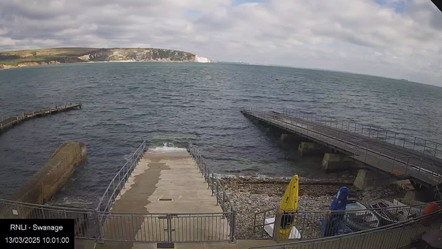 A coastal scene showing a view of the sea with gentle waves. In the foreground, there is a concrete ramp leading to the water, surrounded by a metal railing. On the right side, a small wooden pier extends into the ocean. To the left, there are several boats on the shore, including one yellow kayak. The background features a rocky coastline with cliffs and a cloudy sky overhead. The scene conveys a peaceful harbor atmosphere.