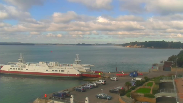 A ferry is docked at a harbor, with a calm sea in the background. The sky is partly cloudy with blue patches peeking through. A few vehicles are parked near the waterfront, and several boats are visible on the water. In the distance, there are green hills and a sandy shore. The setting is tranquil and scenic.