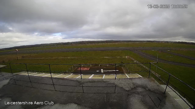 A cloudy sky is visible overhead, with varying shades of gray. Below, an open grass area leads to a dark asphalt runway. The runway is bordered by grass and wooden fences. A small windsock is positioned to the left of the runway. There is a flat area with white markings, possibly indicating a helipad, located in front of a multi-level building with a railing at the edge. The scene is tranquil and appears to be early morning.