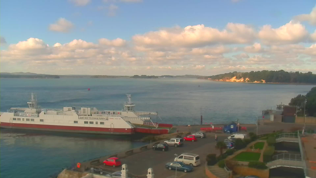 A scenic waterfront view features a large ferry docked at a harbor, surrounded by calm water under a partly cloudy sky. In the background, a green shoreline is visible, with trees lining the coast and a sandy beach. Various vehicles are parked nearby, with some people walking along the promenade. The setting is tranquil, with hints of sunlight illuminating the landscape.