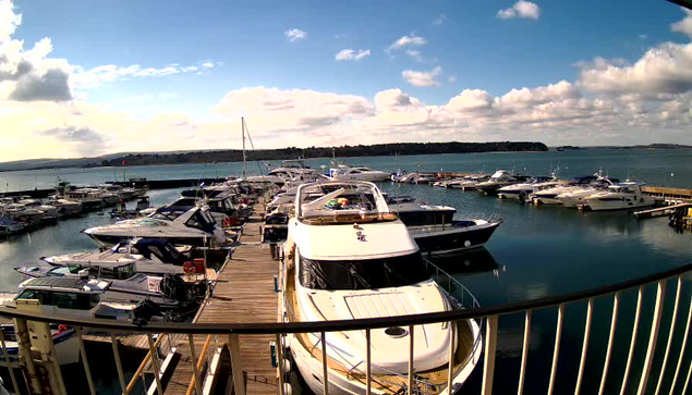 A scenic view of a marina filled with various boats moored along wooden docks. In the foreground, a large white yacht is prominent, with a red and yellow object on its deck. The water is calm, reflecting the sky, which is partly cloudy with patches of blue visible between the clouds. In the background, more boats are docked, and a hillside can be seen on the horizon.