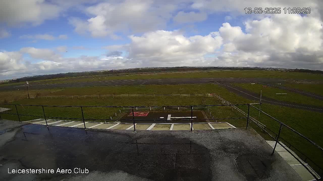 A view from a webcam showing a grassy landscape with a large, open field in the foreground. Parts of a runway are visible, marked by lines and a red area. In the distance, there are several low hills under a partly cloudy sky, with patches of blue peeking through the clouds. There is a railing in the lower part of the image, and the ground appears wet, possibly indicating recent rain. The bottom corner indicates "Leicestershire Aero Club" and a timestamp.