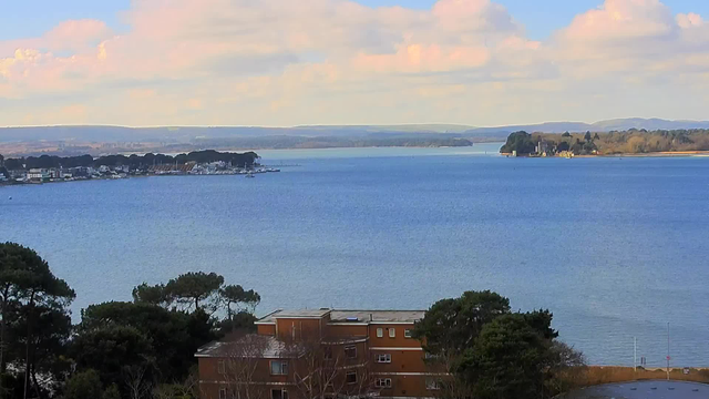 A serene view of a calm body of water, with gentle waves reflecting various shades of blue. In the foreground, there is a brown building partially obscured by trees, which are dark green with some barren branches. To the left, a marina with boats is visible, while in the distance, a shoreline with a mix of trees and buildings stretches along the water's edge. The sky above is bright, with fluffy white clouds scattered across a light blue background, and distant hills are softly outlined on the horizon.