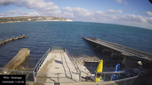 A coastal scene showing clear blue water under a partly cloudy sky. On the left, a concrete pier extends into the water, and to its right, there is a landing area with a ramp leading down. Several boats are visible on the shore, with one yellow kayak and one blue kayak positioned upright on the gravel. In the background, white cliffs rise along the coast, and the horizon meets the ocean in the distance.