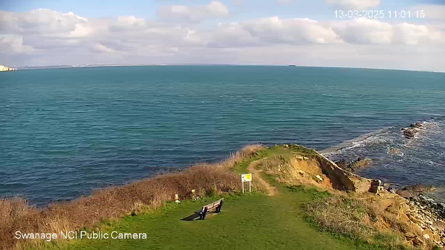 A panoramic view of a coastal scene featuring calm turquoise water under a partly cloudy sky. In the foreground, there is a green grassy area with a wooden bench facing the sea. To the right, rocky cliffs edge the shoreline, where waves gently crash. A warning sign stands near the path leading towards the cliff's edge. The scene captures a tranquil atmosphere with distant ships visible on the horizon.